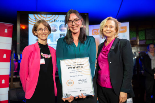 Image of three women smiling at the camera. The woman in the middle is holding a framed certificate that reads 'Fogarty Literary Award Winner'